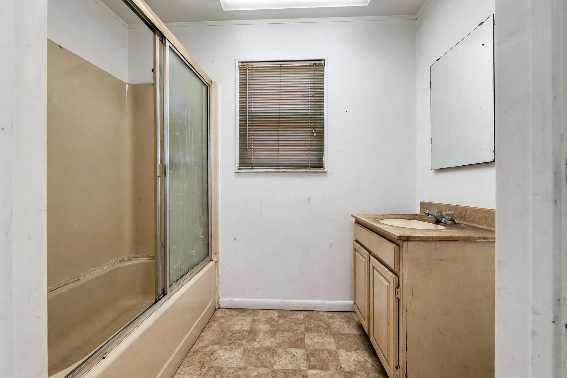 Before: a dated bathroom with a beige tub-shower, worn vanity and vinyl tile floor