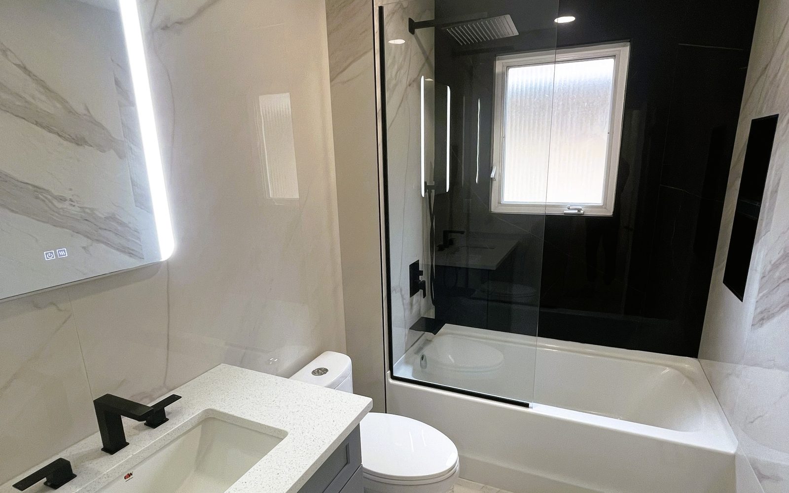 Finished bathroom built by Lions Group: marble-look tile, grey vanity with a backlit mirror and a black glass tub enclosure